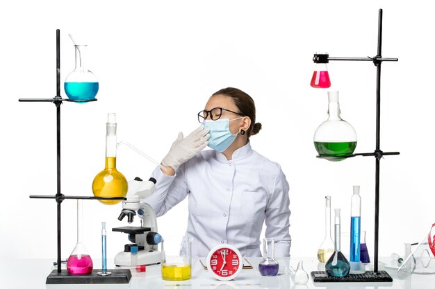 Front view female chemist in medical suit with mask sitting with solutions yawning on white background virus lab chemistry covid splash