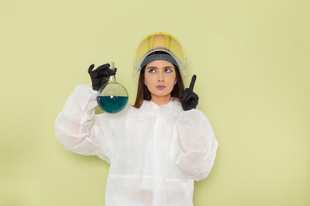 青い溶液でフラスコを保持し、緑の表面で考える特別な防護服の正面図の女性化学者