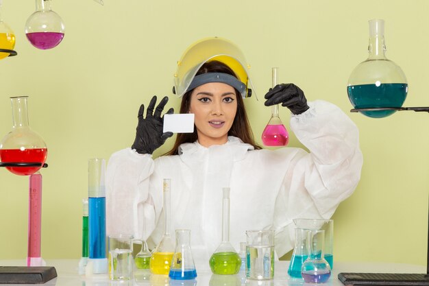 Front view female chemist in special protective suit holding flask with solution and card on light-green surface