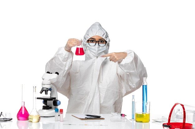 Front view female chemist in special protective suit holding red solution on white background covid- chemistry virus lab