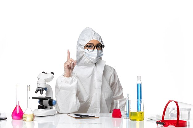 Front view female chemist in special protective suit sitting with solutions on the white background chemistry covid virus pandemic