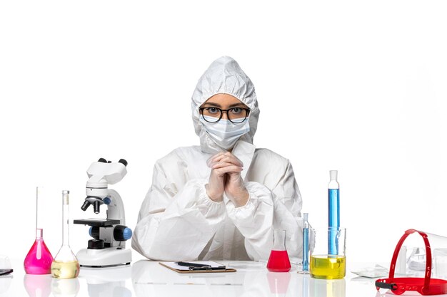 Front view female chemist in special protective suit sitting with solutions on white background chemistry covid virus pandemic