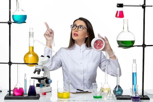 Front view female chemist in white medical suit holding red clocks on a white background science virus lab covid- pandemic