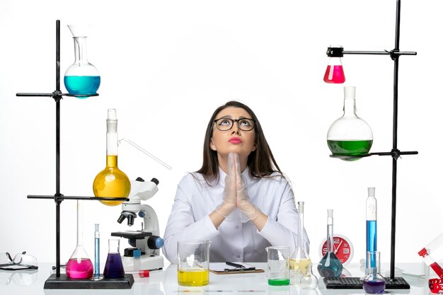Вид спереди женщина-химик в белом медицинском костюме сидит с разными растворами и молится на белом фоне, научный вирус, пандемическая лаборатория covid