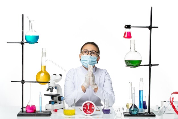 Front view female chemist in white medical suit with mask sitting with solutions on light white background chemist virus covid- splash lab