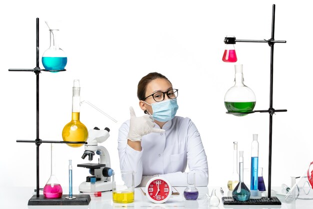 Front view female chemist in white medical suit with mask sitting with solutions on the white background chemist lab virus covid- splash