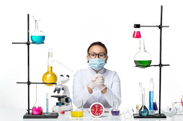 Front view female chemist in white medical suit with mask sitting with solutions on white background chemist virus covid- splash lab