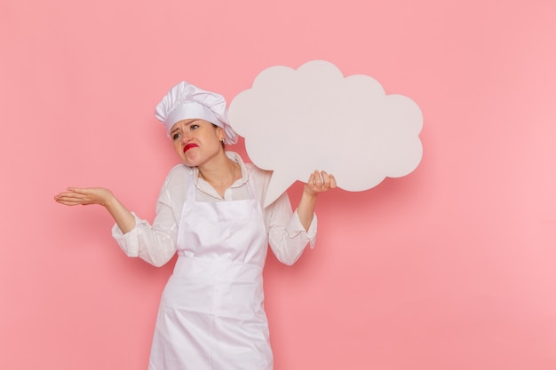 Front view female confectioner in white wear holding a big white sign on the pink wall cook kitchen cuisine food