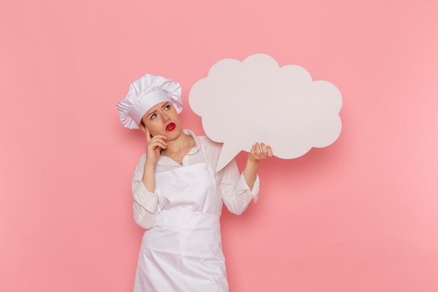 Front view female confectioner in white wear holding a big white sign thinking on the pink wall cook job kitchen cuisine food