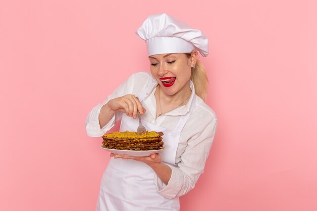 Front view female confectioner in white wear holding delicious pastries designing it on pink wall 