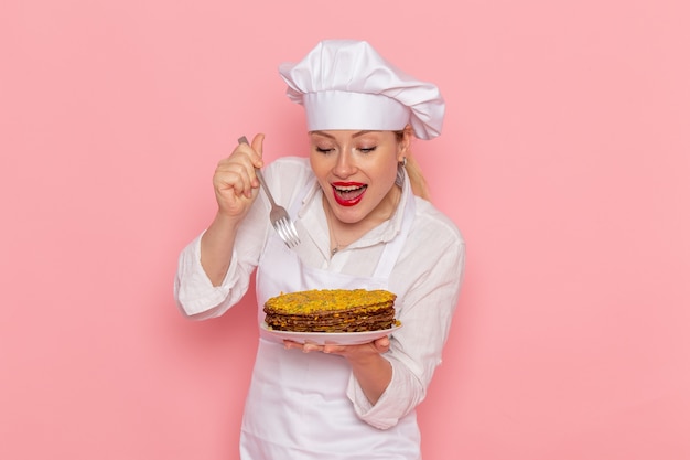 Free photo front view female confectioner in white wear holding delicious pastries and fork on the pink desk