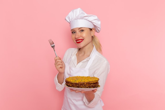 Front view female confectioner in white wear holding delicious pastries and fork on the pink wall 