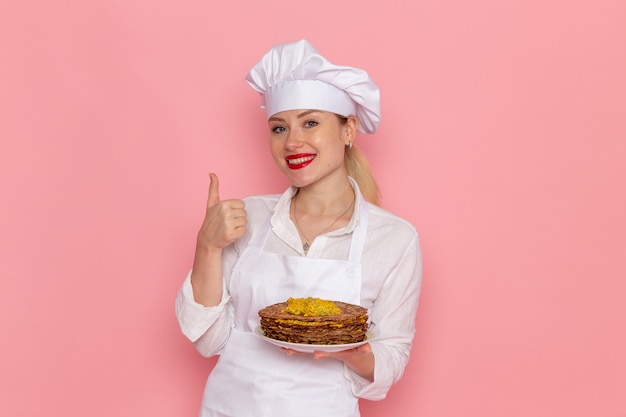 Front view female confectioner in white wear holding delicious pastries on the light pink wall confectionery sweet pastry job work