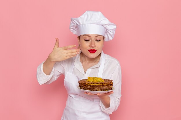 Front view female confectioner in white wear holding delicious pastries on the pink wall confectionery sweet pastry job work