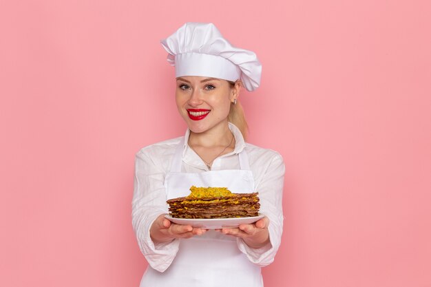 Front view female confectioner in white wear holding delicious pastries smiling on pink wall confectionery sweet pastry job work