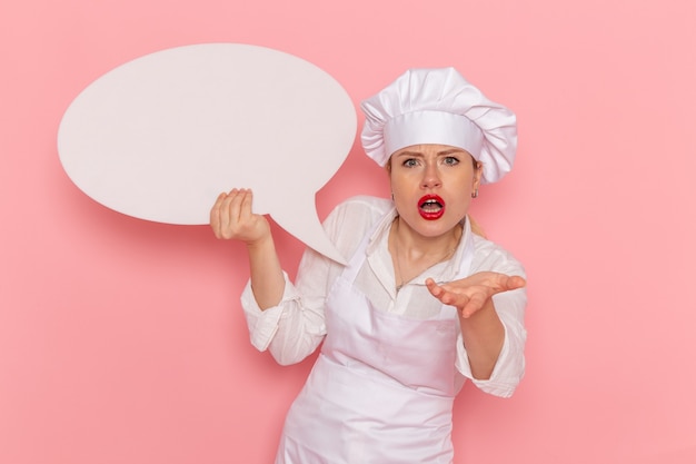 Foto gratuita pasticcere femminile di vista frontale in usura bianca che tiene segno bianco sul lavoro di lavoro della pasticceria dolce della pasticceria della parete rosa