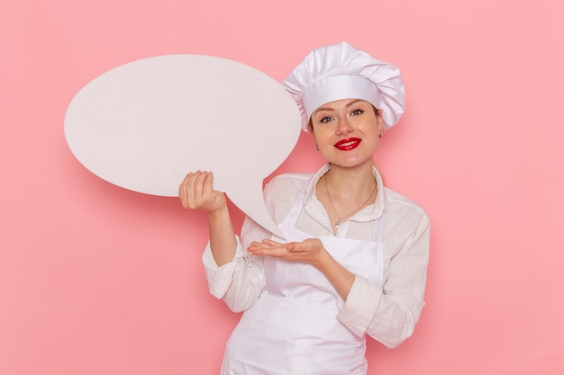 白い看板を保持し、ピンクの机の菓子に笑みを浮かべて白い服を着た女性菓子職人の正面図甘いペストリーの仕事