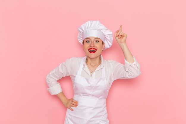 Front view female confectioner in white wear posing with delighted expression on light pink wall confectionery sweet pastry job work