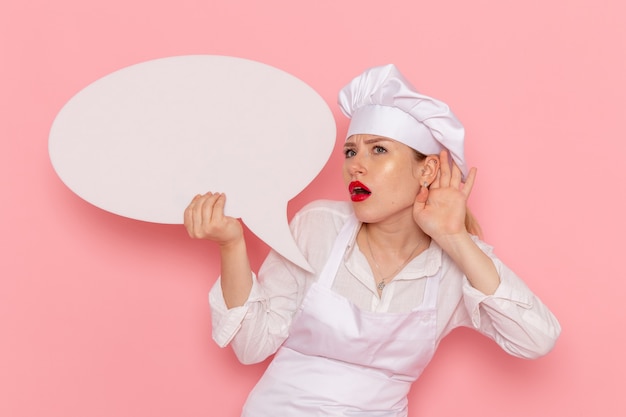 Free photo front view female confectioner in white wear posing with white sign trying to hear out on pink wall confectionery sweet pastry job work