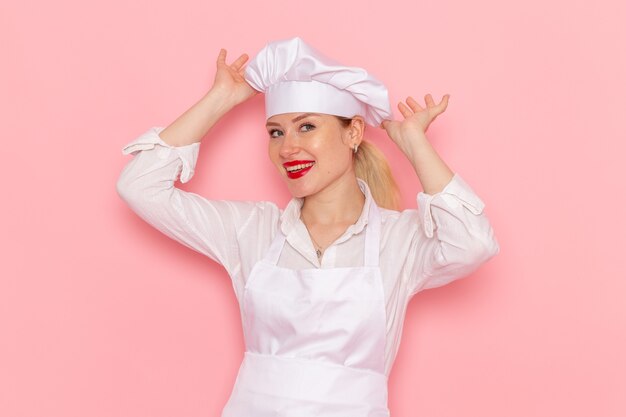 Front view female confectioner in white wear smiling and posing on light pink desk confectionery sweet pastry job work