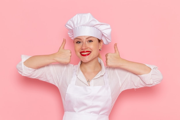Front view female confectioner in white wear smiling and posing on the pink wall cook confectionery sweet pastry work