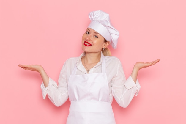 Front view female confectioner in white wear smiling and posing on the pink wall cook confectionery sweet pastry