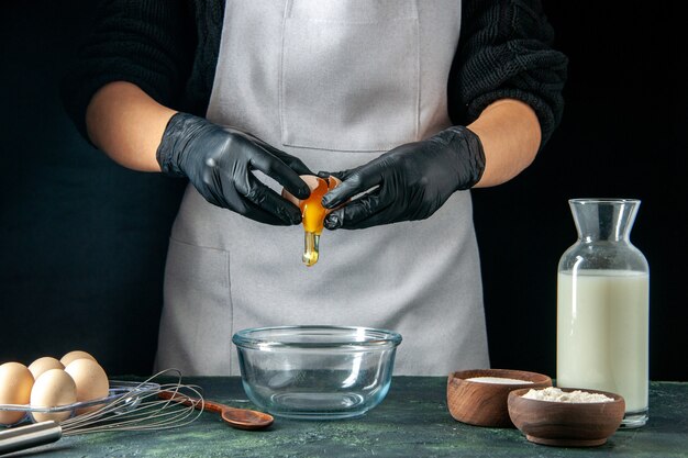 Front view female cook breaking eggs for dough on a dark pastry job cake pies bakery worker cuisine