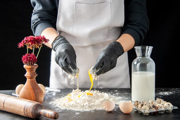 Вид спереди женщина-повар, разбивающая яйца в муку на темной работе, выпечка пирога, выпечки торта, бисквитного теста, выпечки