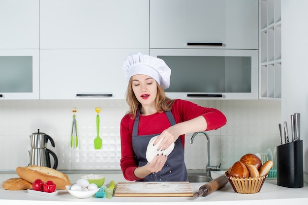 Front view female cook in cook hat holding up dough in the kitchen