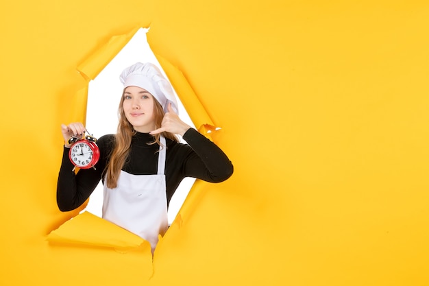 Бесплатное фото Вид спереди женщина-повар в белой кепке повара держит часы на желтом фото, цветная работа, кухня, солнце, еда, эмоции