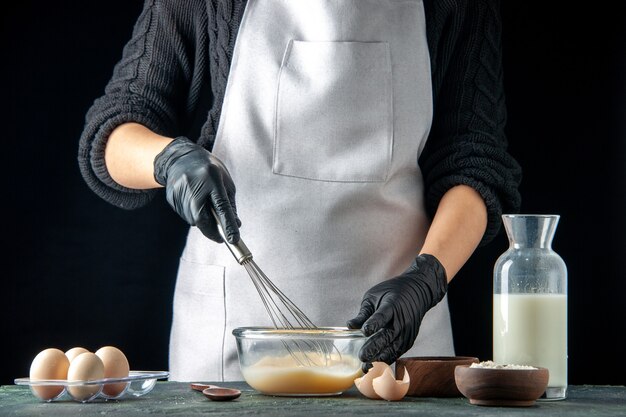 전면 보기 여성 요리사는 어두운 페이스트리 케이크 파이 작업자 반죽 요리 작업 핫케이크에 반죽을 위해 계란과 설탕을 섞습니다.