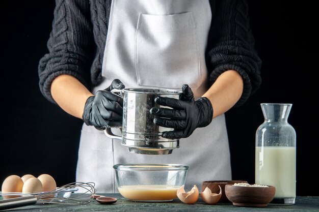 Front view female cook pouring flour into plate with eggs for dough on dark pastry cake pie worker cuisine job hotcake dough