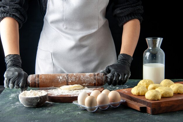 Front view female cook rolling out dough with flour on the dark cake job oven hotcake dough bake pie worker egg cuisine