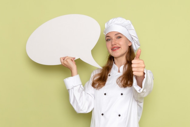 Front view of female cook wearing white cook suit holding big sign on green desk kitchen cuisine cooking female color