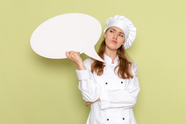 Front view of female cook wearing white cook suit holding white sign and thinking on light-green wall