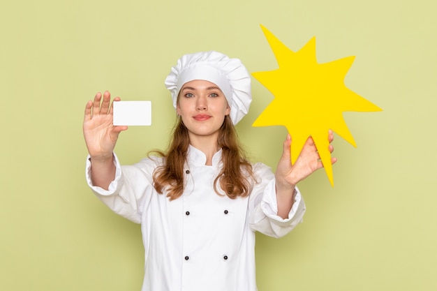 Front view of female cook wearing white cook suit holding yellow sign and card on green wall