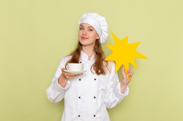 Front view of female cook wearing white cook suit holding yellow sign with cup of coffee on green desk kitchen cuisine cooking meal female color