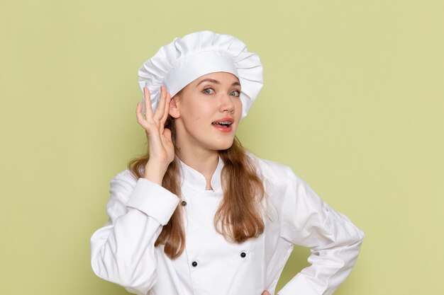 緑の壁に耳を傾けようとしている白いクックスーツを着ている女性料理人の正面図