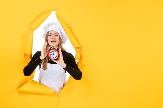 Front view female cook in white cook cap holding clock on yellow photo color job cuisine kitchen sun emotion