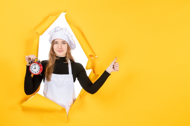 Front view female cook in white cook cap holding clock on yellow photo color job cuisine kitchen sun food emotion