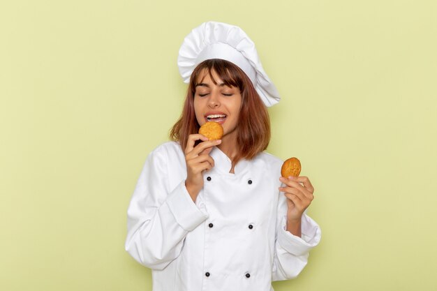 Front view female cook in white cook suit eating cookies on green surface