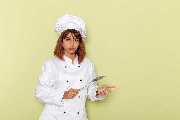 Front view female cook in white cook suit holding big silver spoon on the green surface