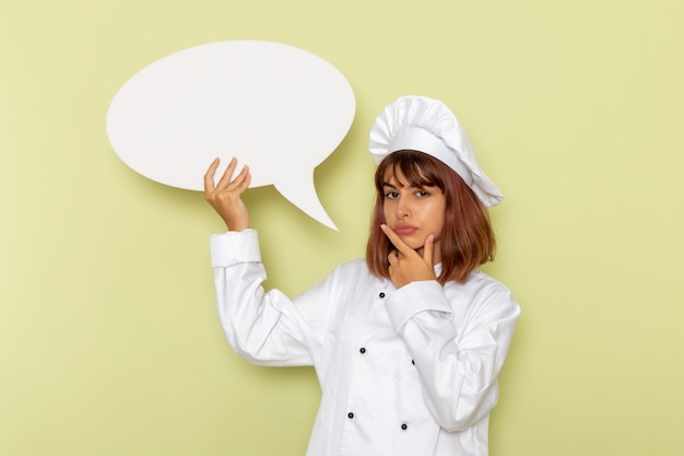 Front view female cook in white cook suit holding a big white sign on green surface