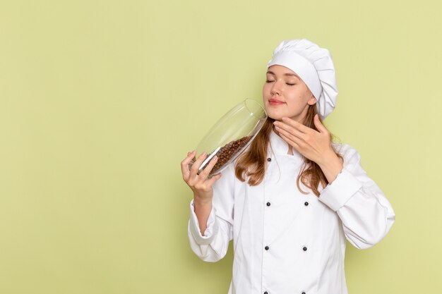 Front view of female cook in white cook suit holding can with brown coffee seeds on green wall