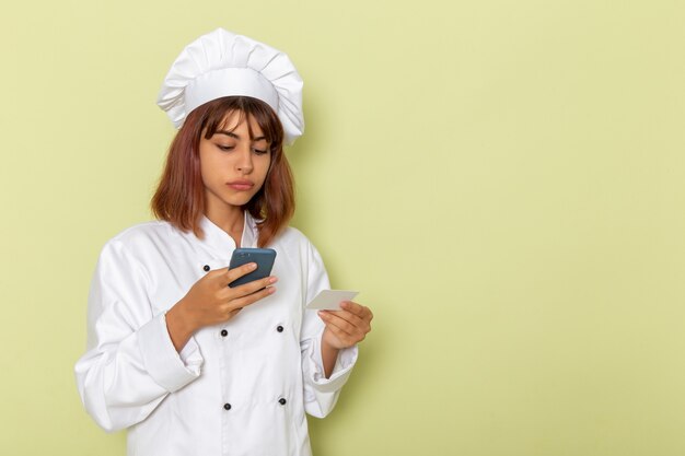 Женщина-повар, вид спереди в белом костюме повара, держит карту и смартфон на зеленом столе