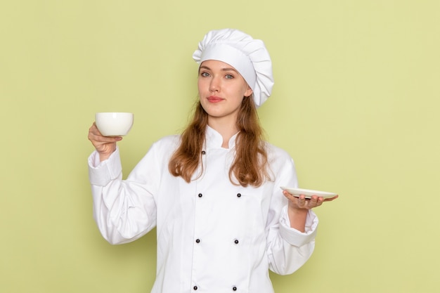 Front view of female cook in white cook suit holding cup and plate on green wall