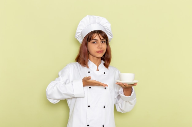 Front view female cook in white cook suit holding cup of tea on a green surface