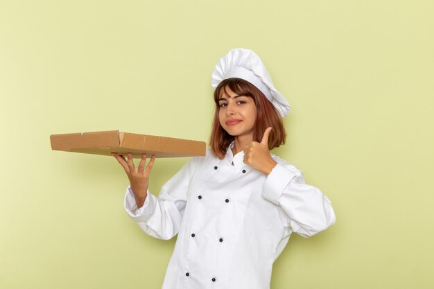 Женщина-повар, вид спереди в белом костюме повара, держа коробку для еды на зеленой поверхности