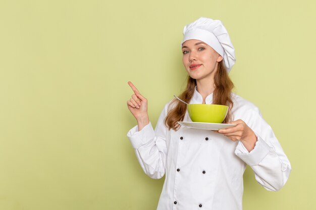 Front view of female cook in white cook suit holding green plate on the light green wall