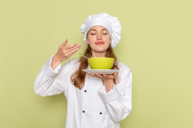 Front view of female cook in white cook suit holding green plate and smelling on green wall